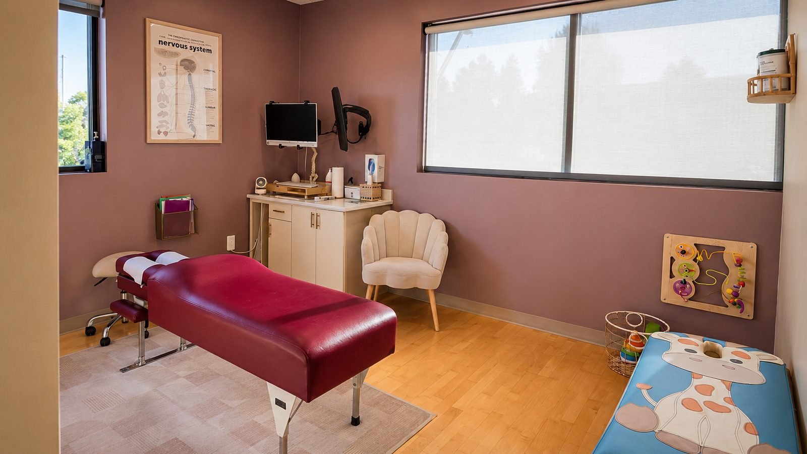 Treatment room at Empower Family Chiropractic with a burgundy adjusting table, a giraffe-print pediatric adjusting table, and a nervous system chart on the wall
